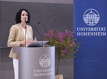 Eine Frau steht in einem Auditorium an einem Pult und hält eine Rede.