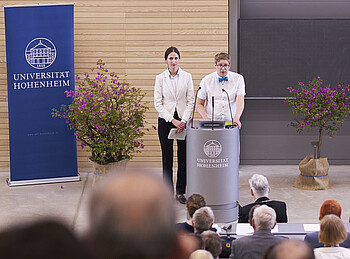 Zwei Personen stehen in einem Auditorium an einem Pult und sprechen zu den Menschen in den Sitzreihen.