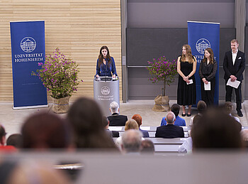 In einem Auditorium hält eine Frau an einem Podium eine Rede. Drei weitere Personen stehen in kleinem Abstand zu ihr ebenfalls vor den zuhörenden Menschen.