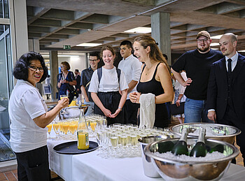 An einem festlichen Tisch schenkt eine Frau Saft und Sekt aus. Viele Menschen stehen vor dem Tisch und freuen sich auf ihr Getränk.