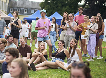 Viele junge Menschen stehen und sitzen auf einer Wiese. Sie schauen alle in dieselbe Richtung und freuen sich.