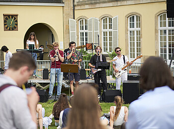 Sechs Menschen spielen Gitarre, Keyboard und Drums. Zwei Personen davon singen. Eine Gruppe Menschen schaut ihnen zu.