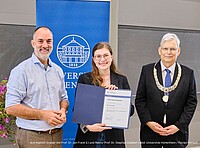 Drei Menschen stehen beieinander und freuen sich über den Preis, den einer der Menschen verliehen bekommen hat.