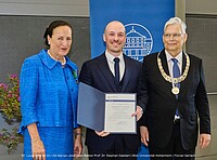 Drei Menschen stehen beieinander und freuen sich über den Preis, den einer der Menschen verliehen bekommen hat.